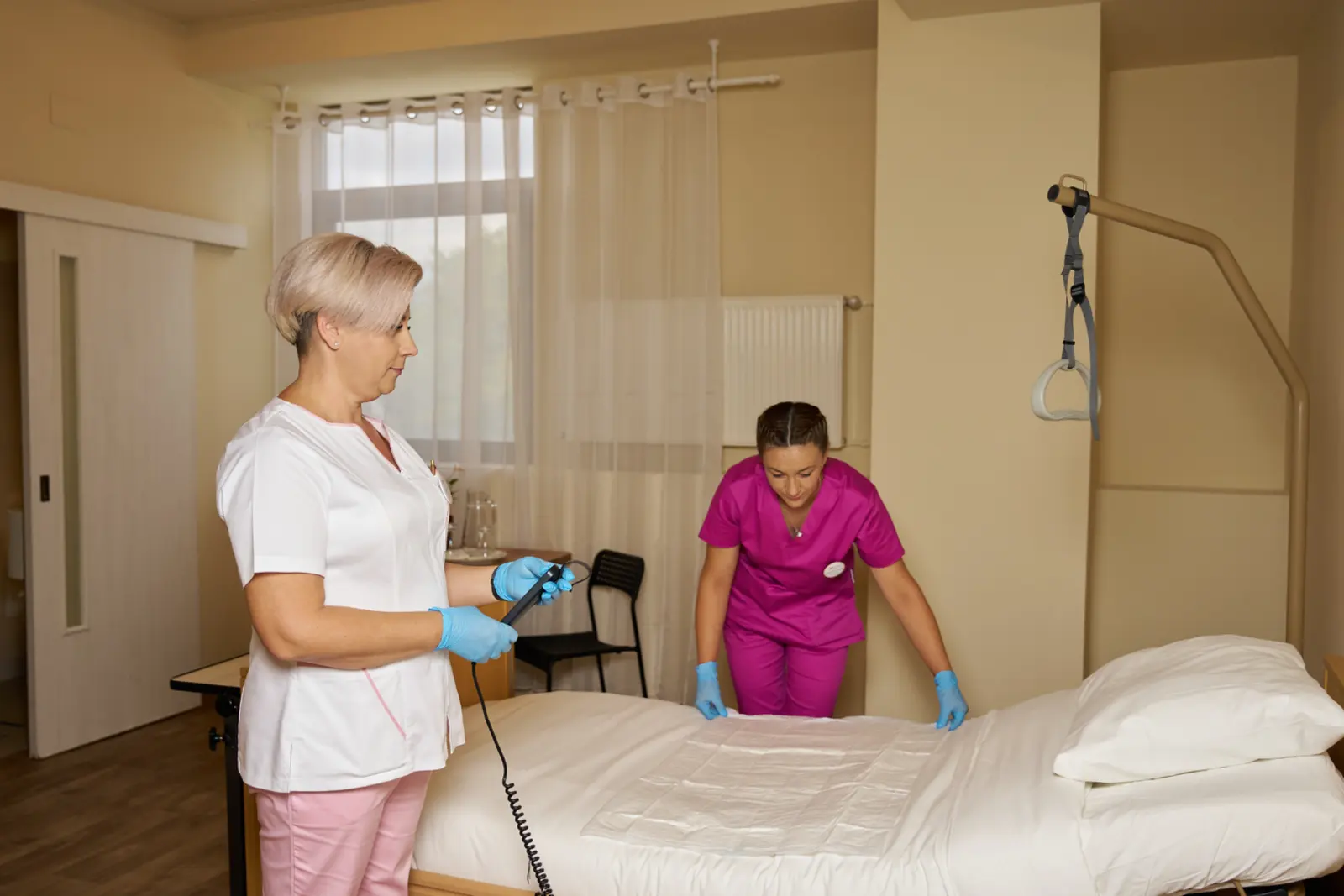 Două asistente medicale în uniforme albe și roz, cu mănuși albastre, pregătesc salteaua, AsertivO Otopeni