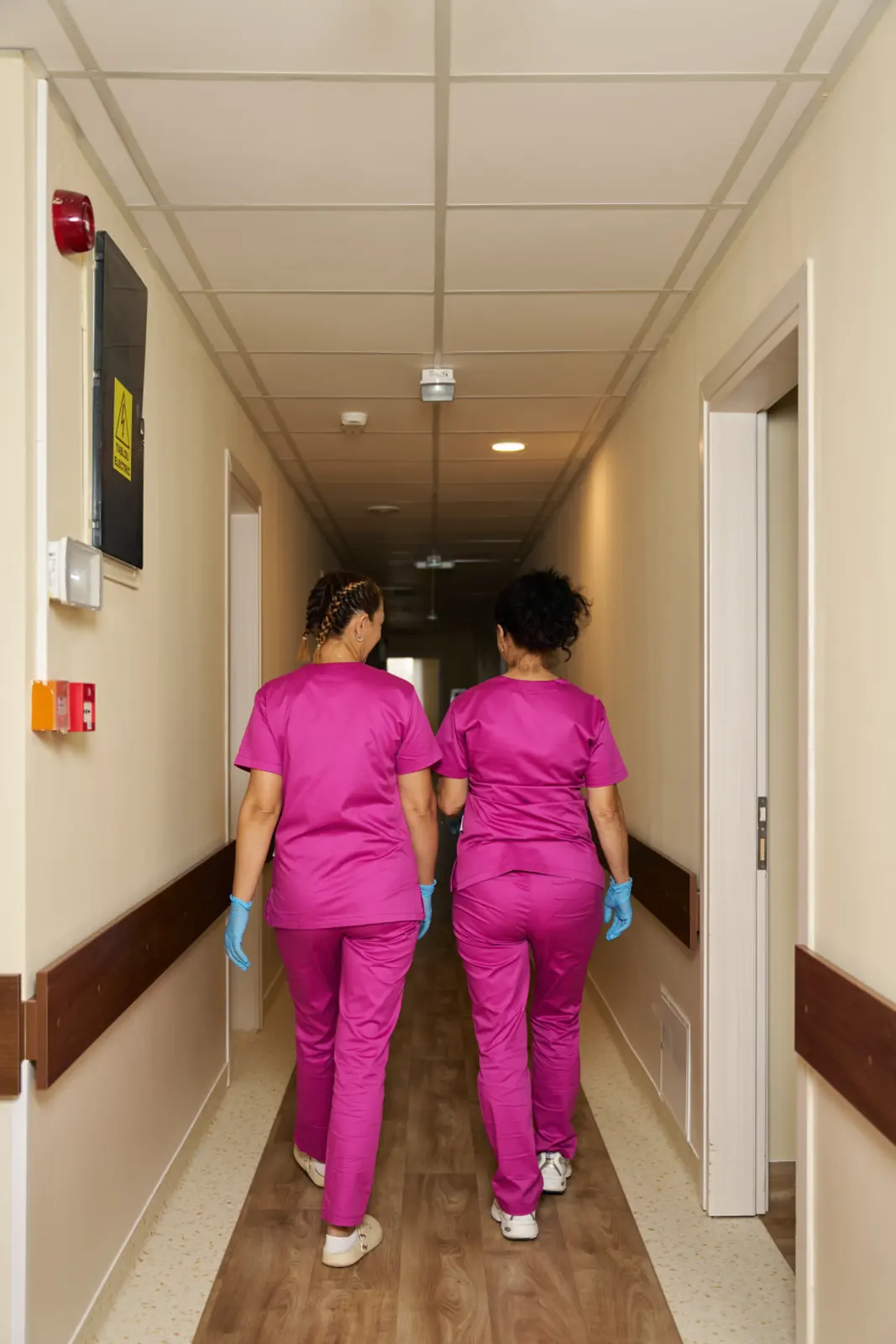 Două cadre medicale îmbrăcate în halate roz și mănuși albastre de protecție mergând pe coridorul, AsertivO Otopeni