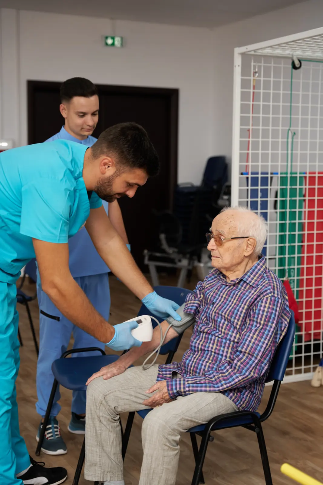 Două cadre medicale în scruburi turcoaz măsoară tensiunea arterială unui senior cu ochelari, AsertivO Otopeni