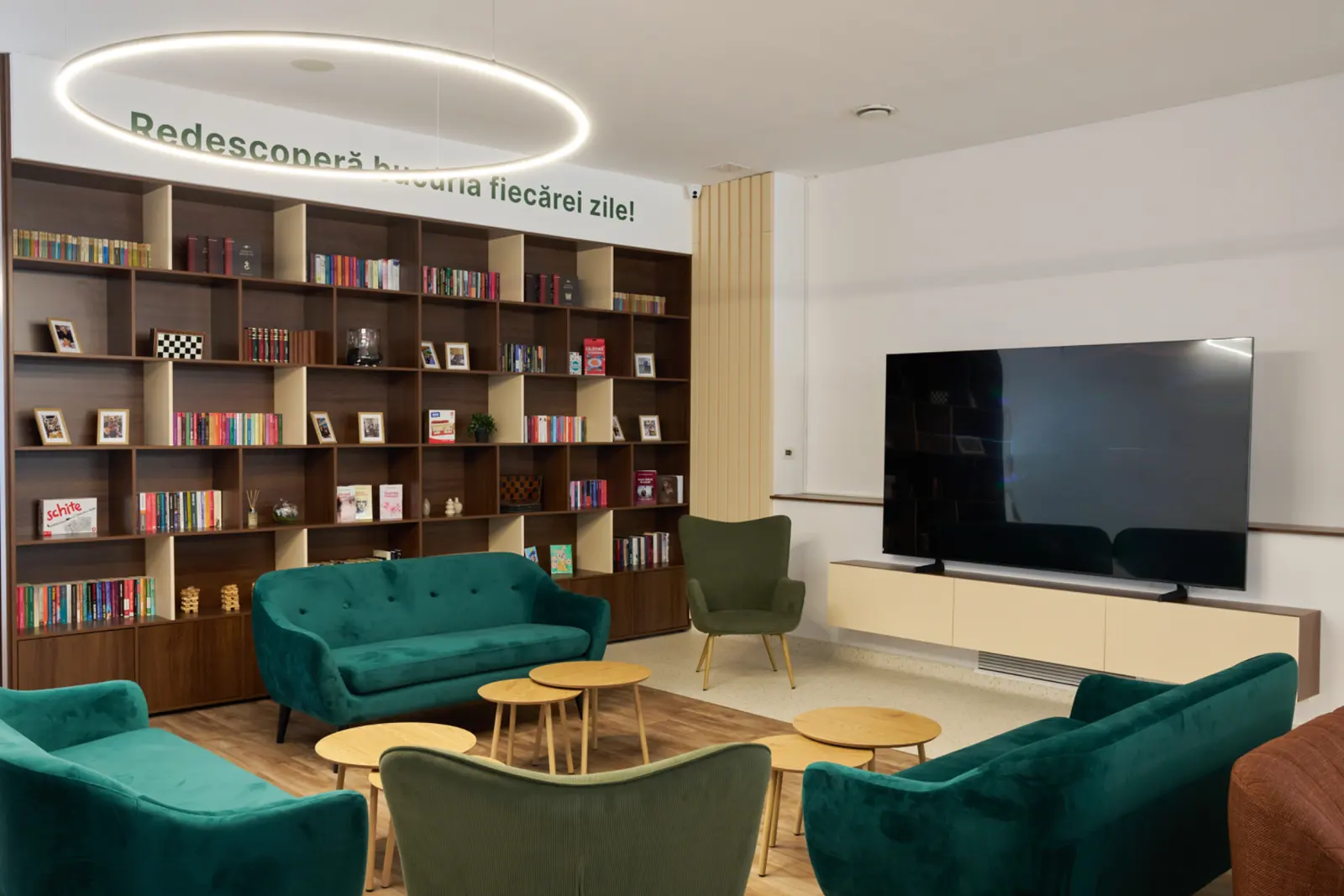 Spațiu luminos cu bibliotecă plină de cărți, canapele de catifea verde, măsuțe din lemn și televizor mare, AsertivO Otopeni